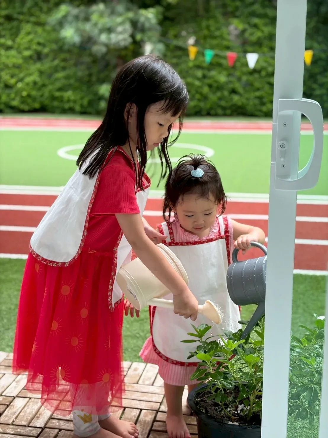 An older girl steadying the watering can for a younger one at the plant pot