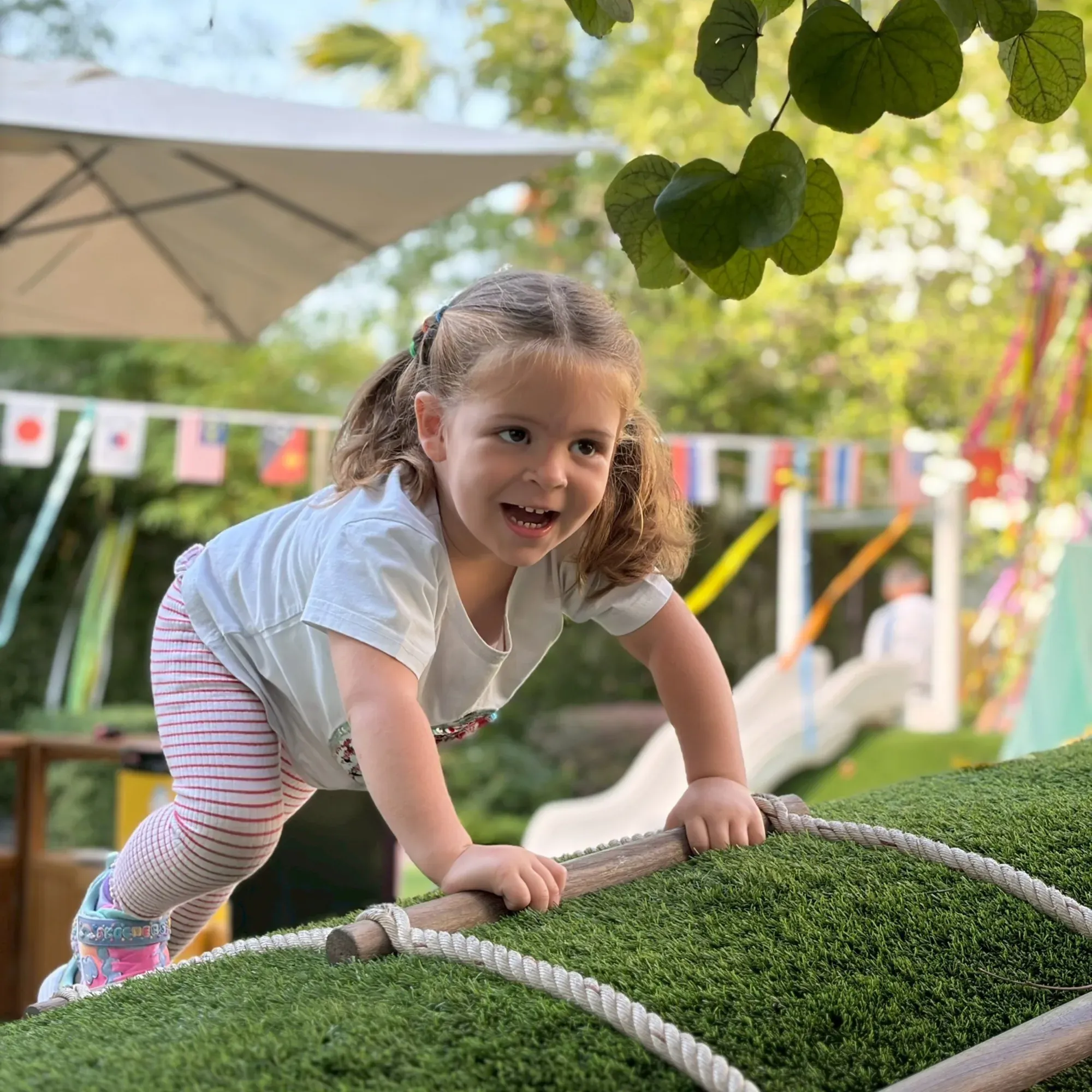 A girl laughing as she climbs a grassy rope hill in the Creative Play Space, with flags and a slide behind her