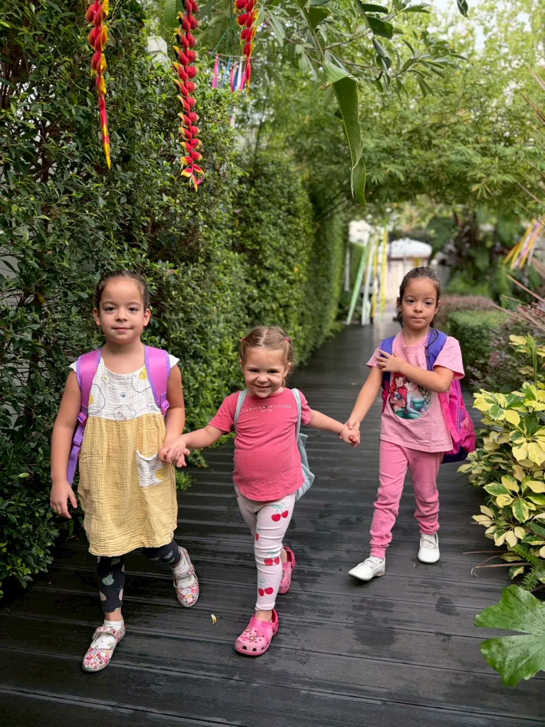 Three children with their backpacks walking hand in hand along the garden path into school