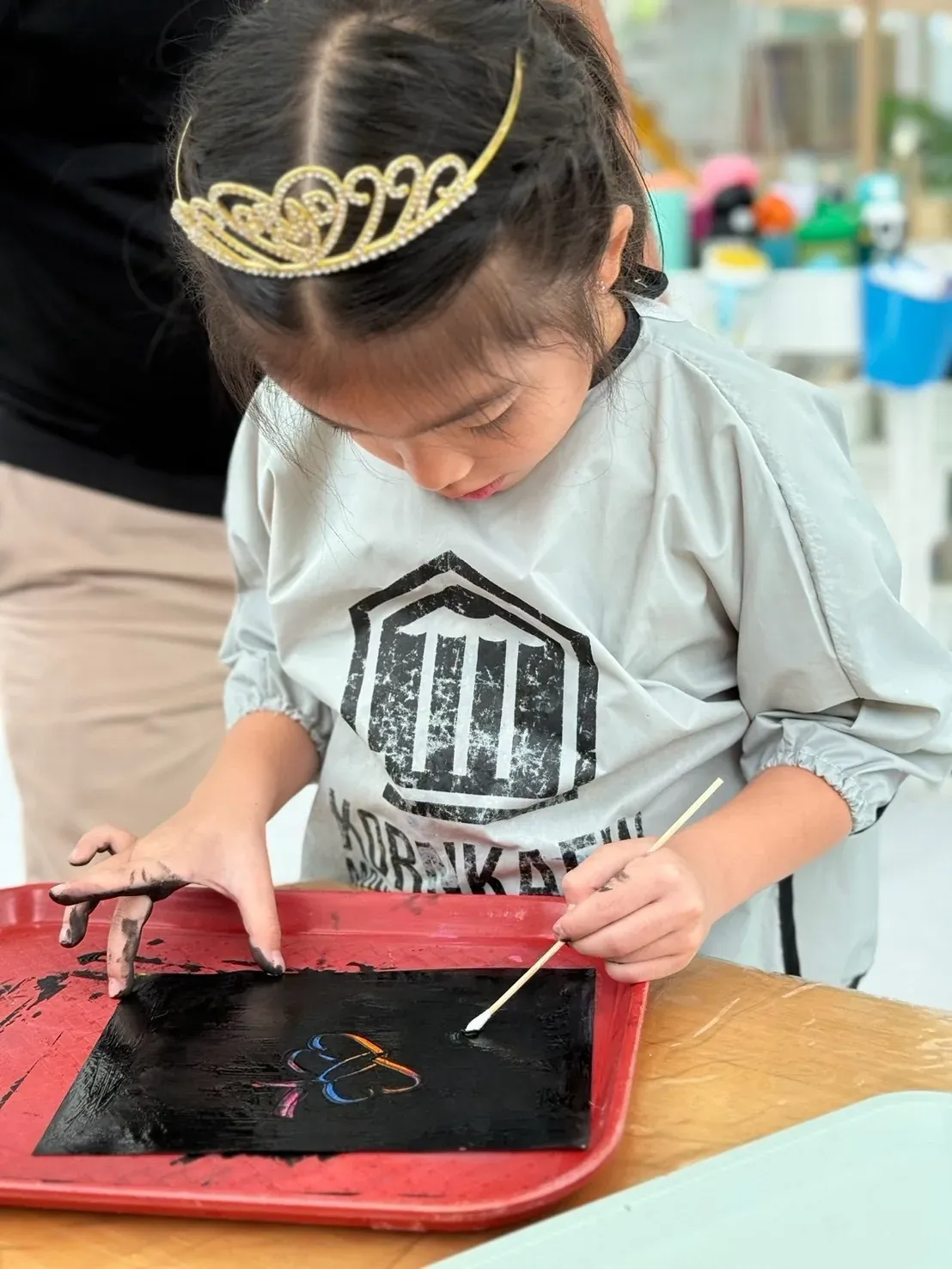 A girl in a tiara scratching a rainbow flower out of black scratch paper