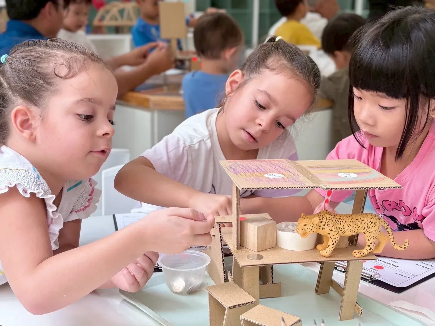 Three girls testing the cardboard shelter they built for a toy leopard