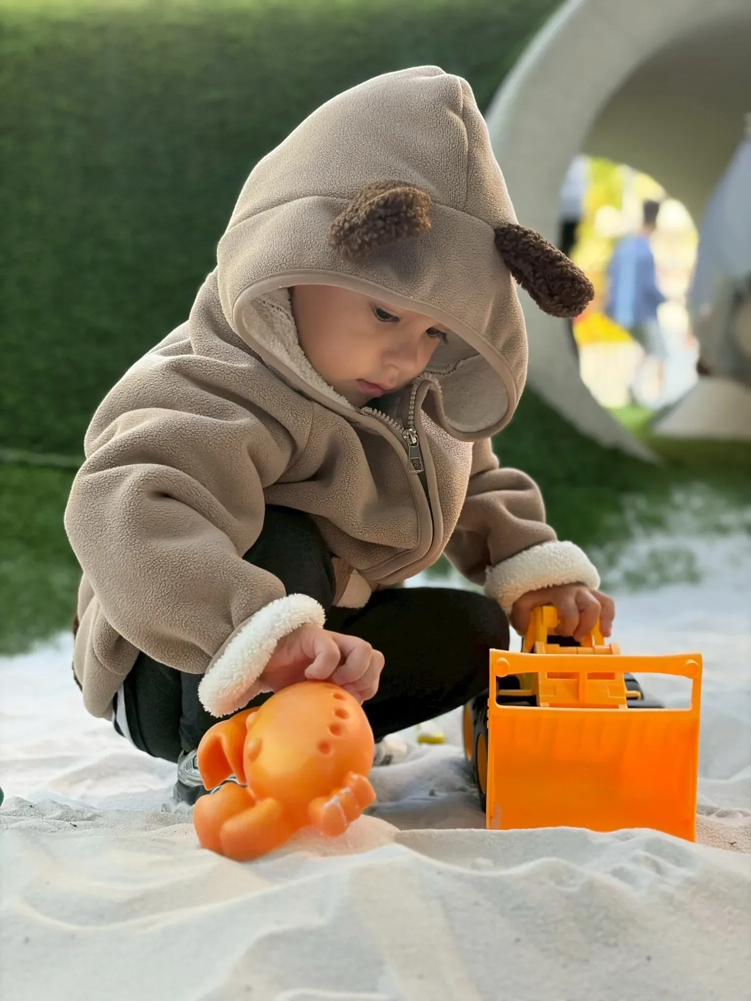 A toddler in a bear-ear hood playing with a digger in the sand