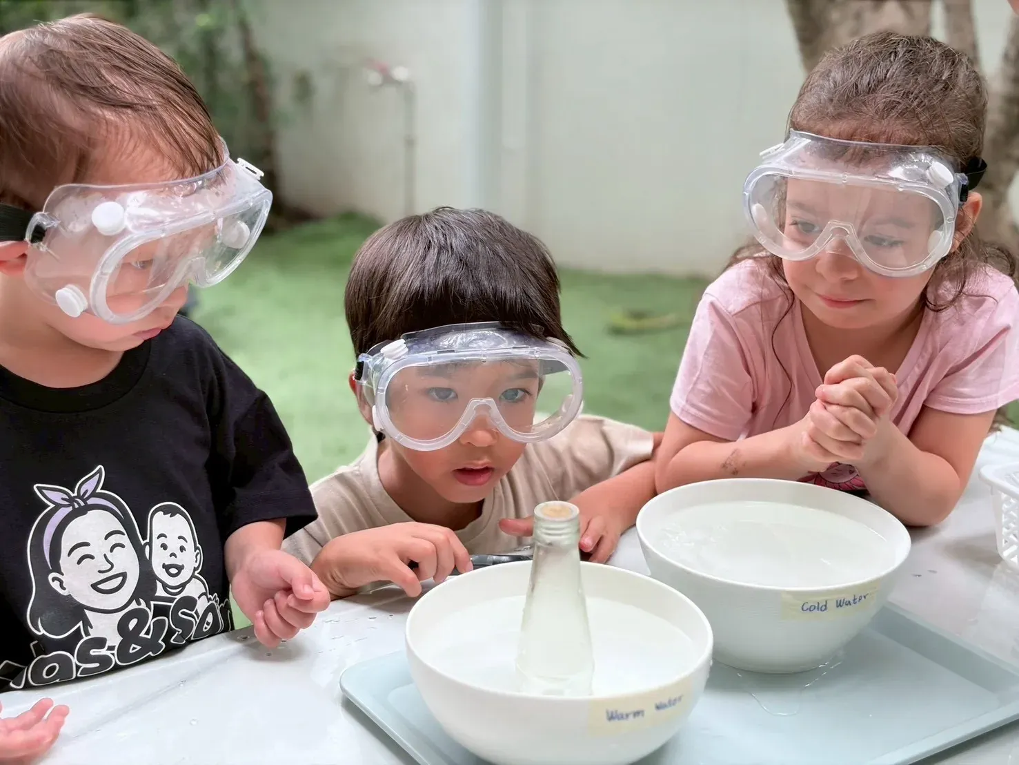 Three children in safety goggles watching a bottle in bowls of warm and cold water