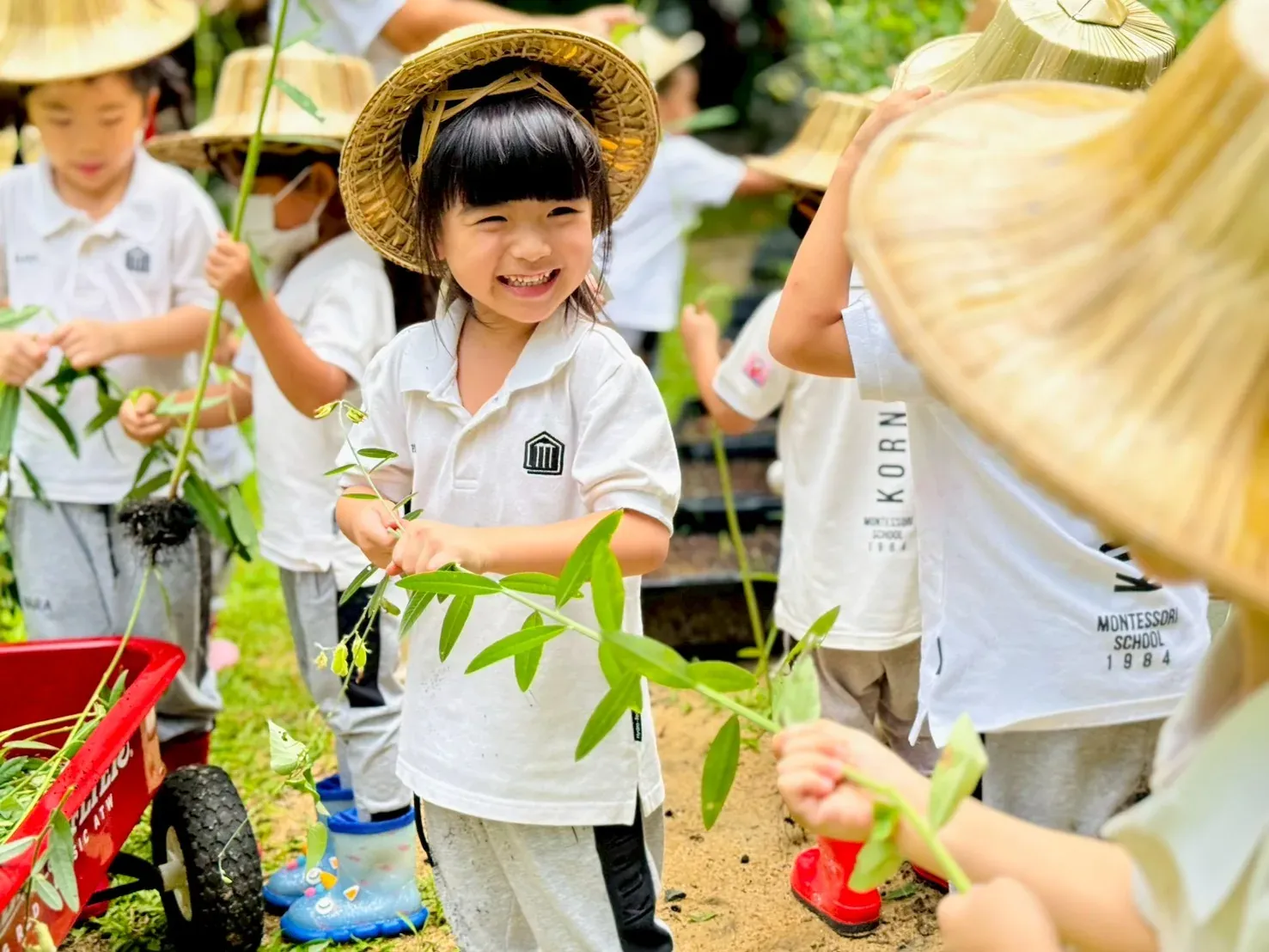 A smiling Kornkaew girl in a straw hat holding a seedling, with classmates planting in the school garden at the Dusit campus