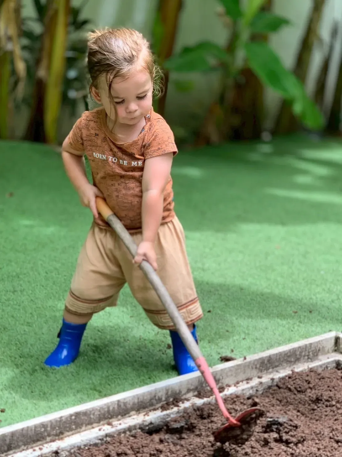 A toddler in blue boots turning the soil of a raised bed with a full-size hoe