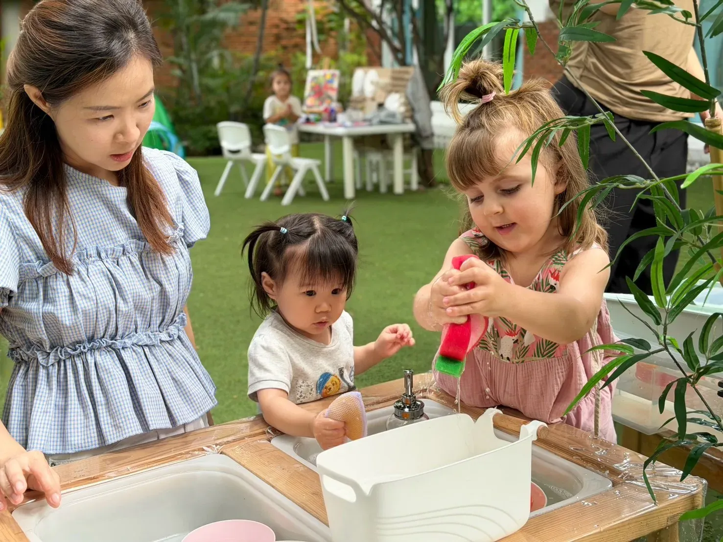 Two children washing up with sponges at a water play station, with a parent watching